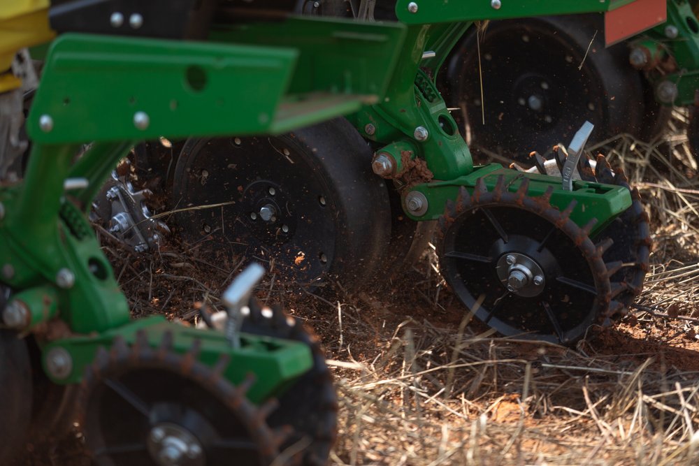 A close-up view of a no-till planter in action, planting seeds into the soil with minimal disturbance to the ground. The machine's disc openers and seed tubes are visible, ensuring precise seed placement